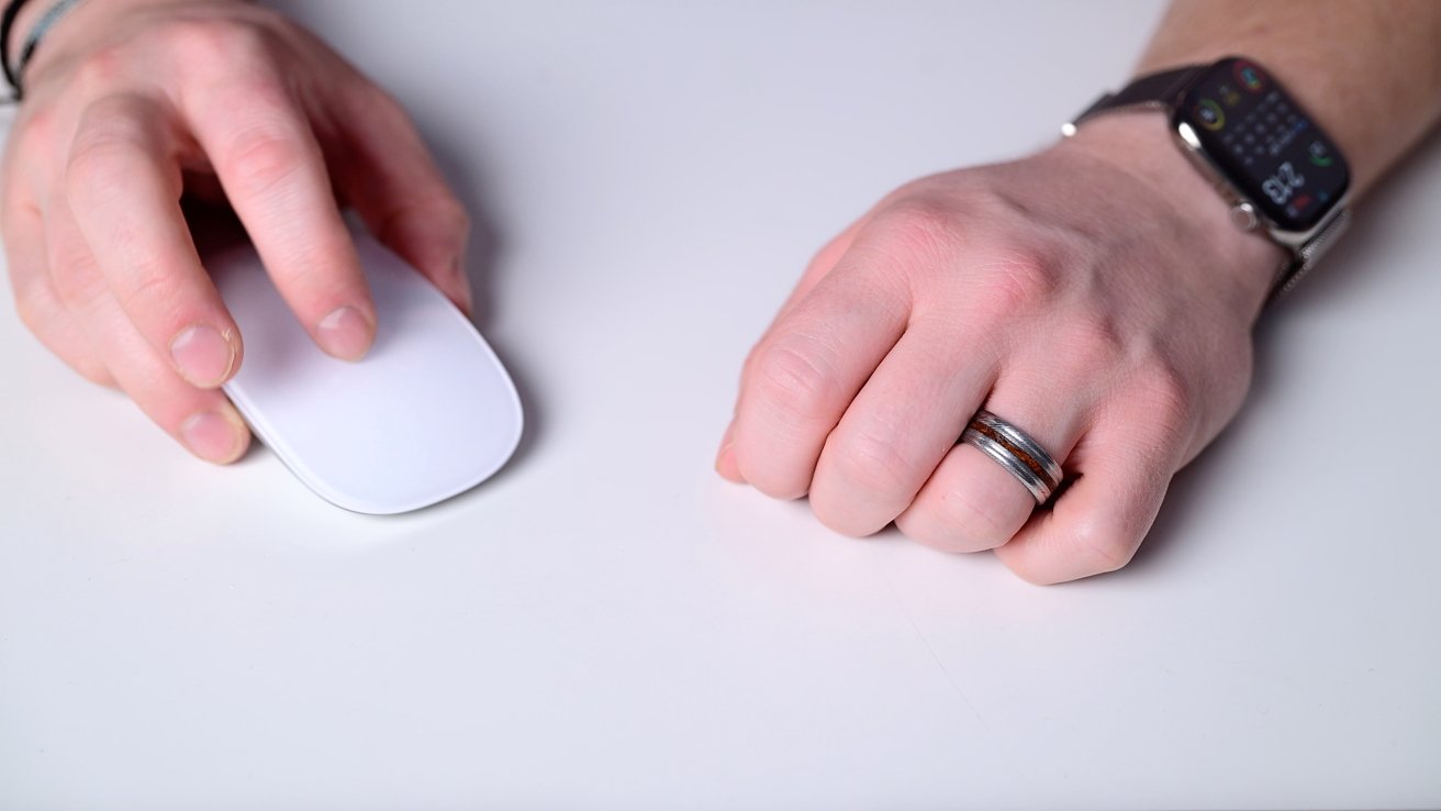 Two hands on a white surface, one holding a computer mouse, the other wearing a ring and watch.