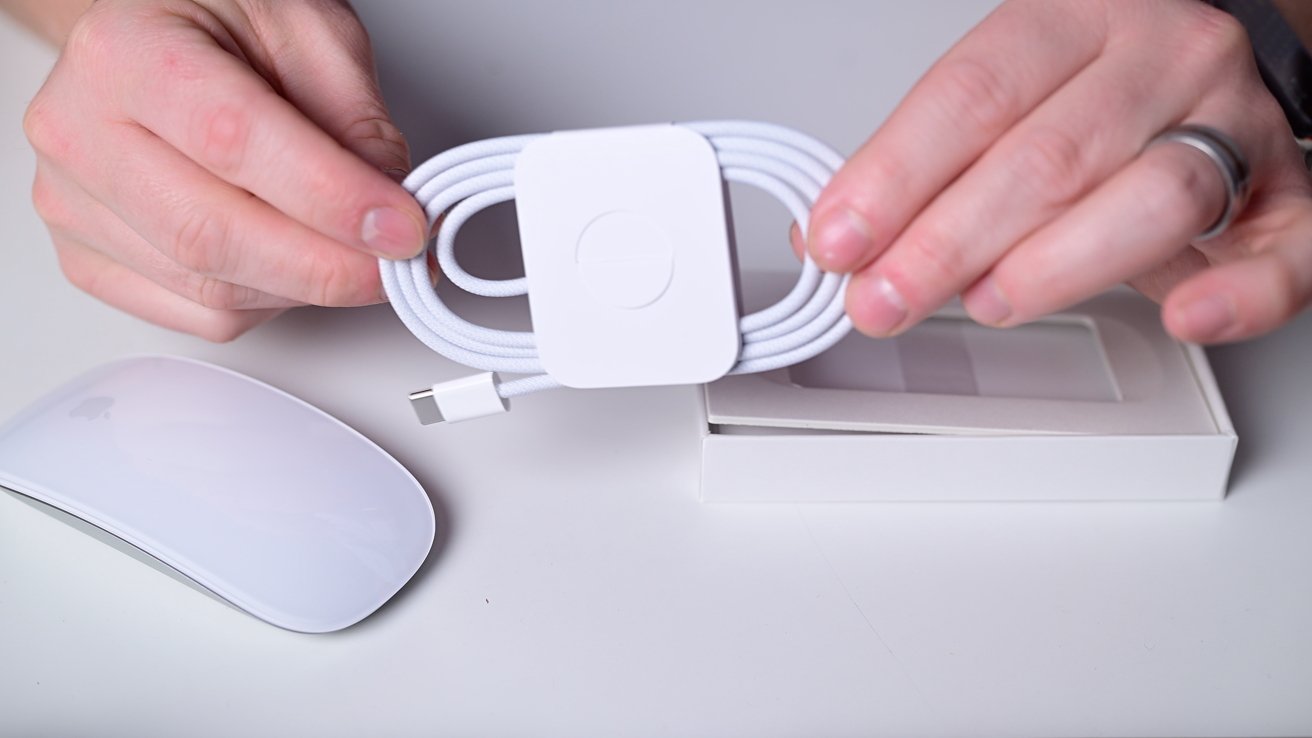 Hands holding a coiled white USB-C cable above a white box, next to a sleek white computer mouse on a white surface.