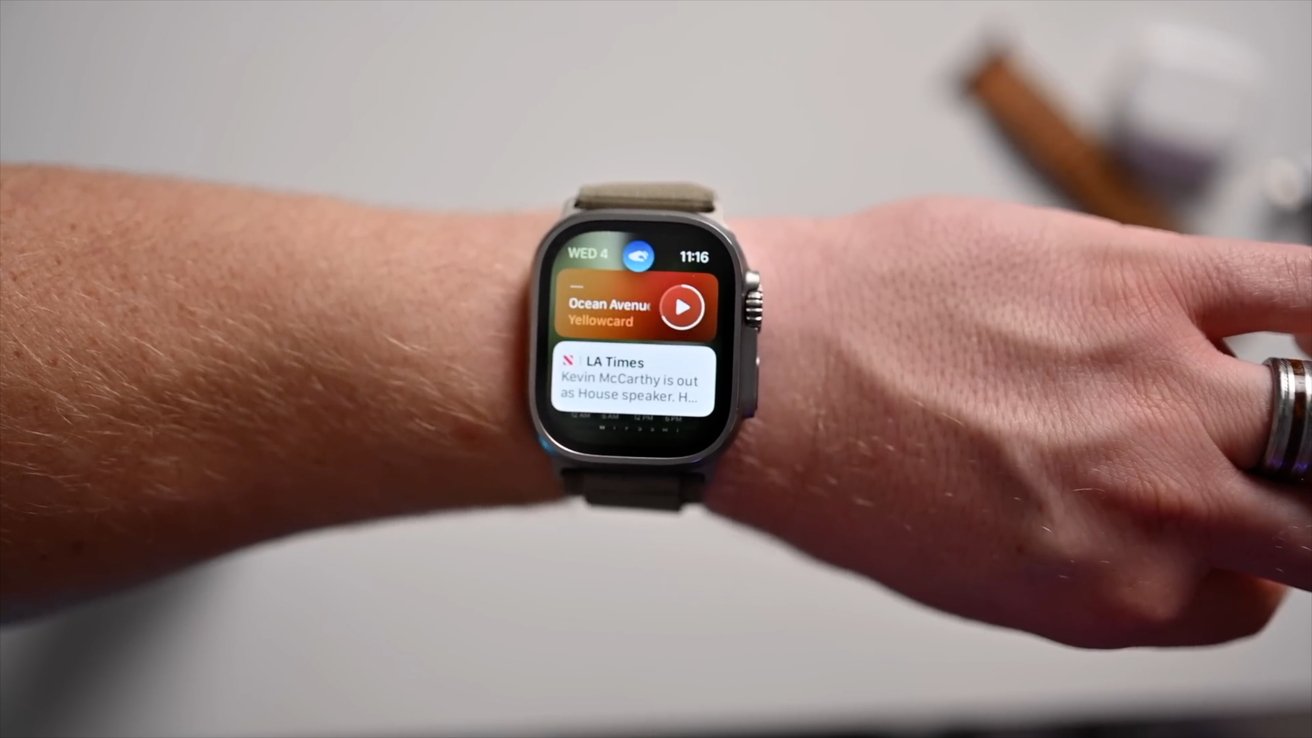 A person wearing a smartwatch displaying music player controls and a news notification on their wrist.