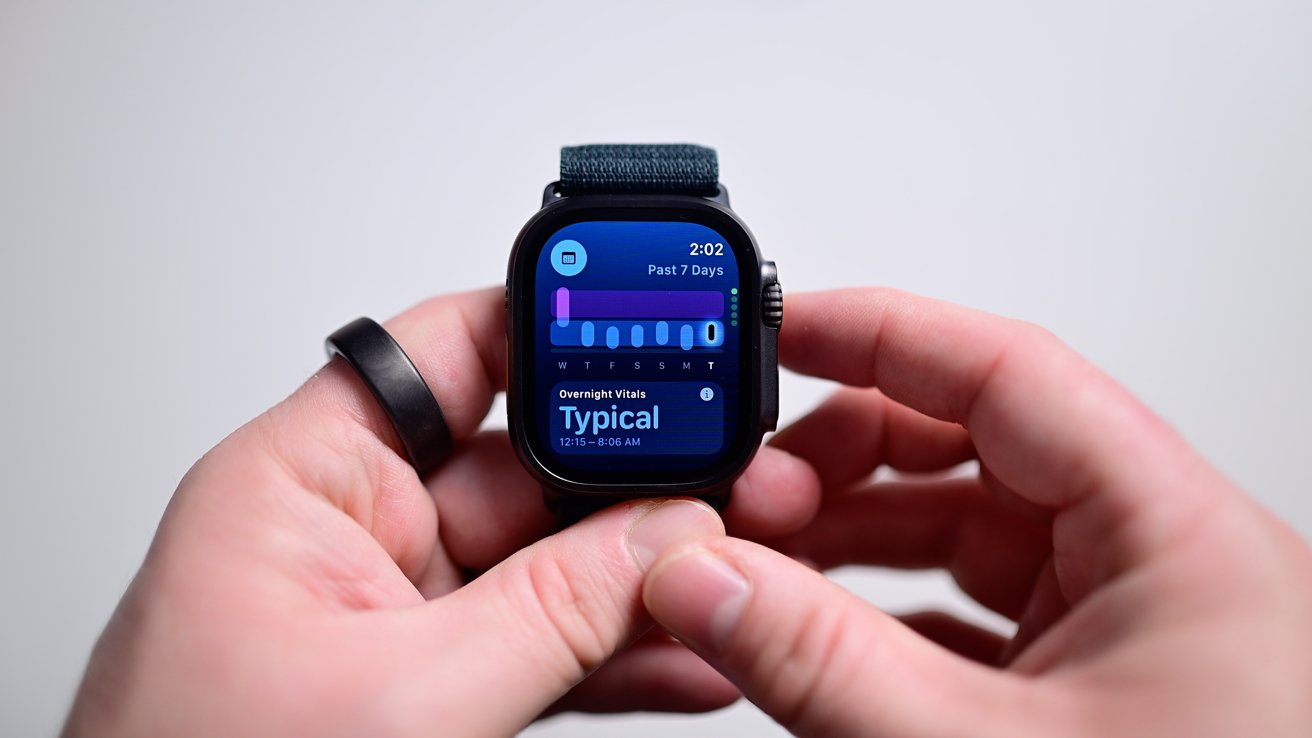 Hand holding a smartwatch displaying health data, with graphs and text: 'Overnight Vitals Typical,' showing information from the past week.