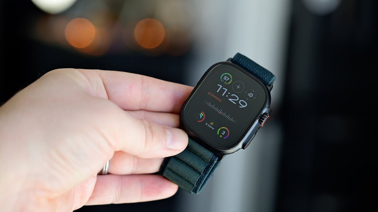 Hand holding a modern smartwatch with a dark screen, displaying the time 11:29, various icons, and a green fabric strap.