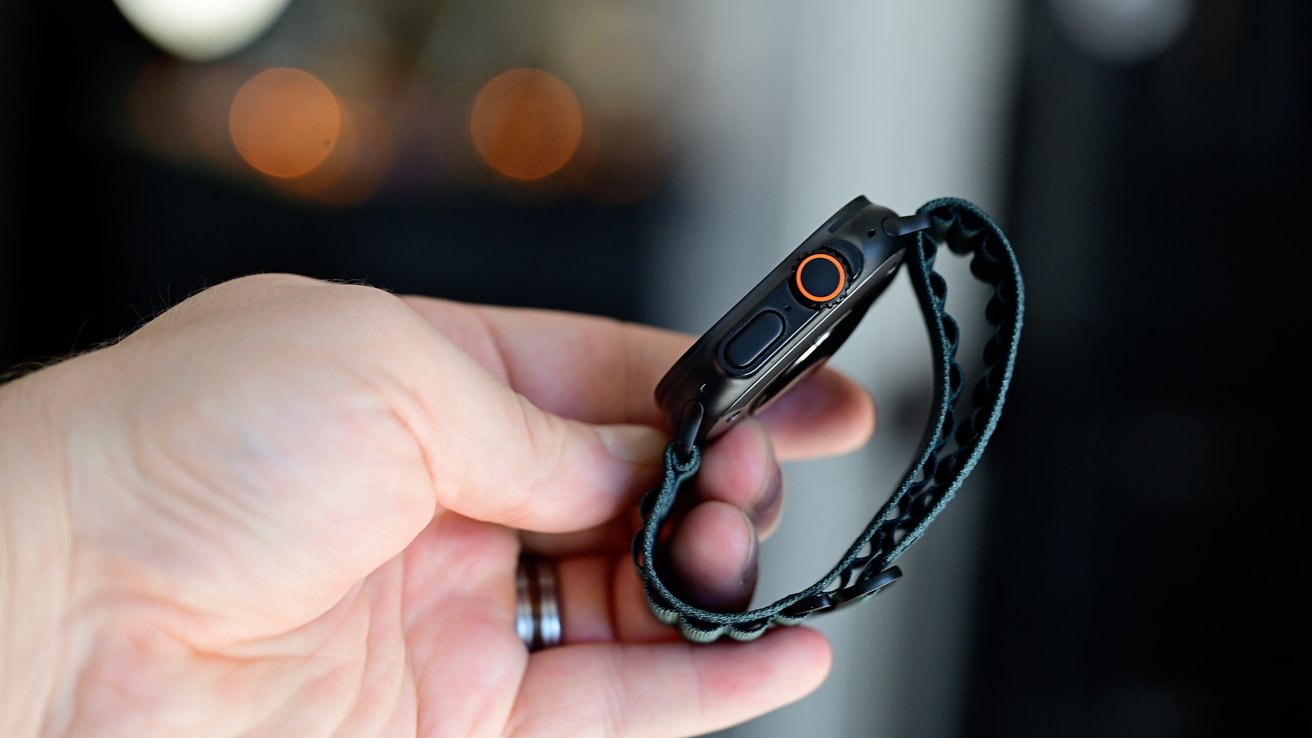 A hand holding a wristwatch with a black case, orange button, and woven green strap, blurred background.