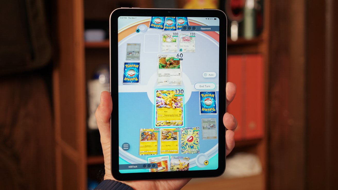Hand holding a tablet displaying a Pokemon trading card game with various colorful cards on screen. Bookshelf in the blurred background.