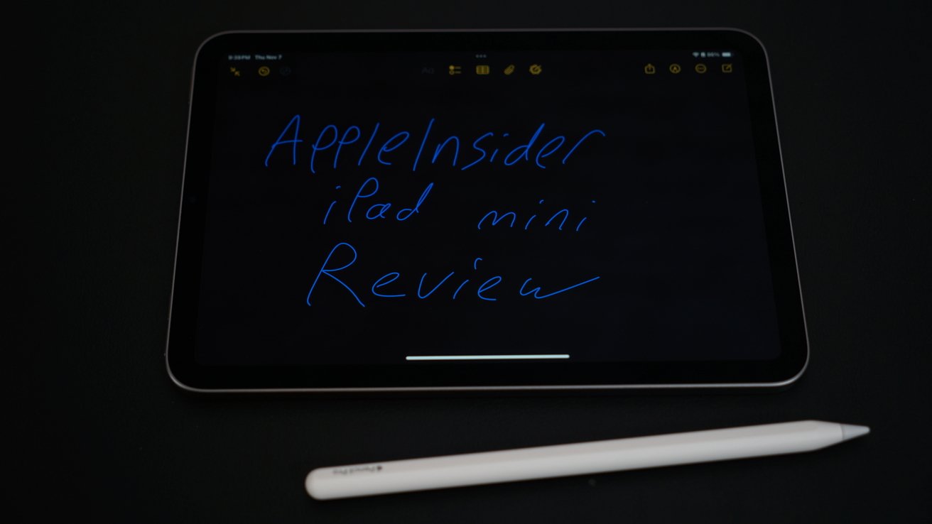 iPad with handwritten text 'AppleInsider iPad mini Review' on screen, placed on a dark surface with a white stylus beside it.