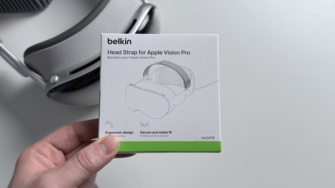 Hand holding Belkin head strap box for Apple Vision Pro, with a sketch of the product and text highlighting ergonomic design, secure fit. VR headset in the background.