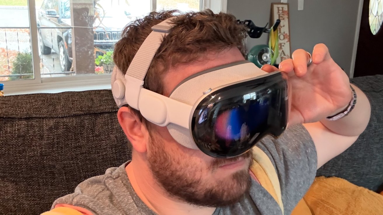 A person is wearing a virtual reality headset while sitting on a couch with a window and parked car visible in the background.