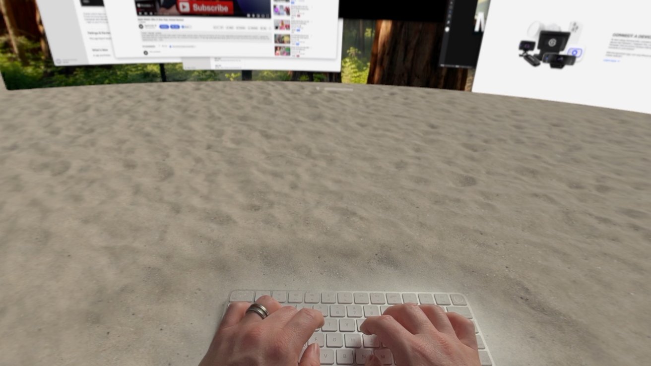 Hands typing on a keyboard placed on sand, with large computer screens displaying webpages in the background.