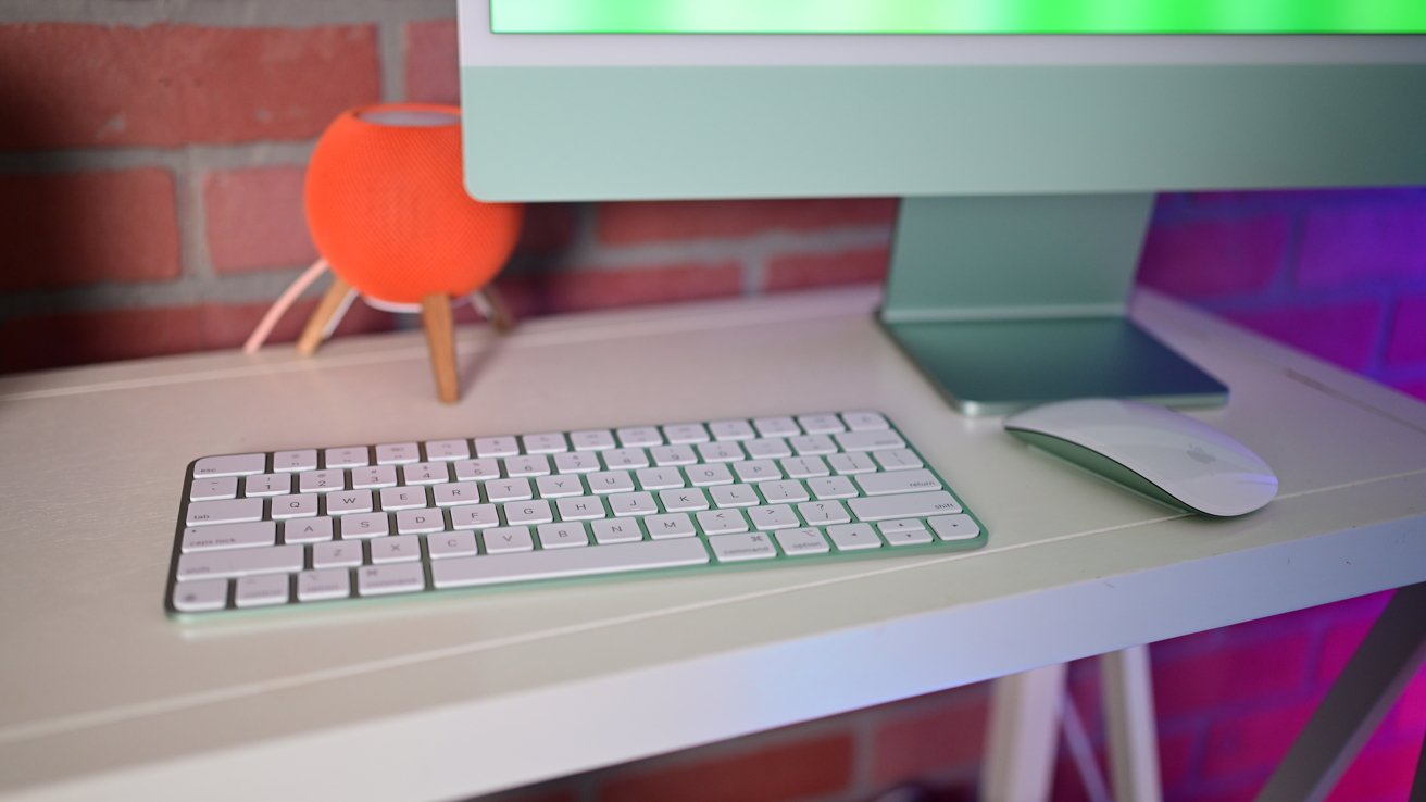 Modern computer setup with a green monitor, white keyboard, and mouse on a white desk. An orange spherical speaker stands against a red brick wall.