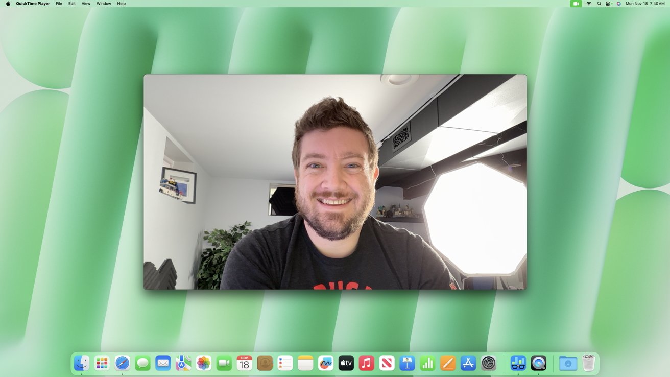 Smiling man with short hair in a room with plants and a bright light, displayed on a computer screen with a green background and app icons.