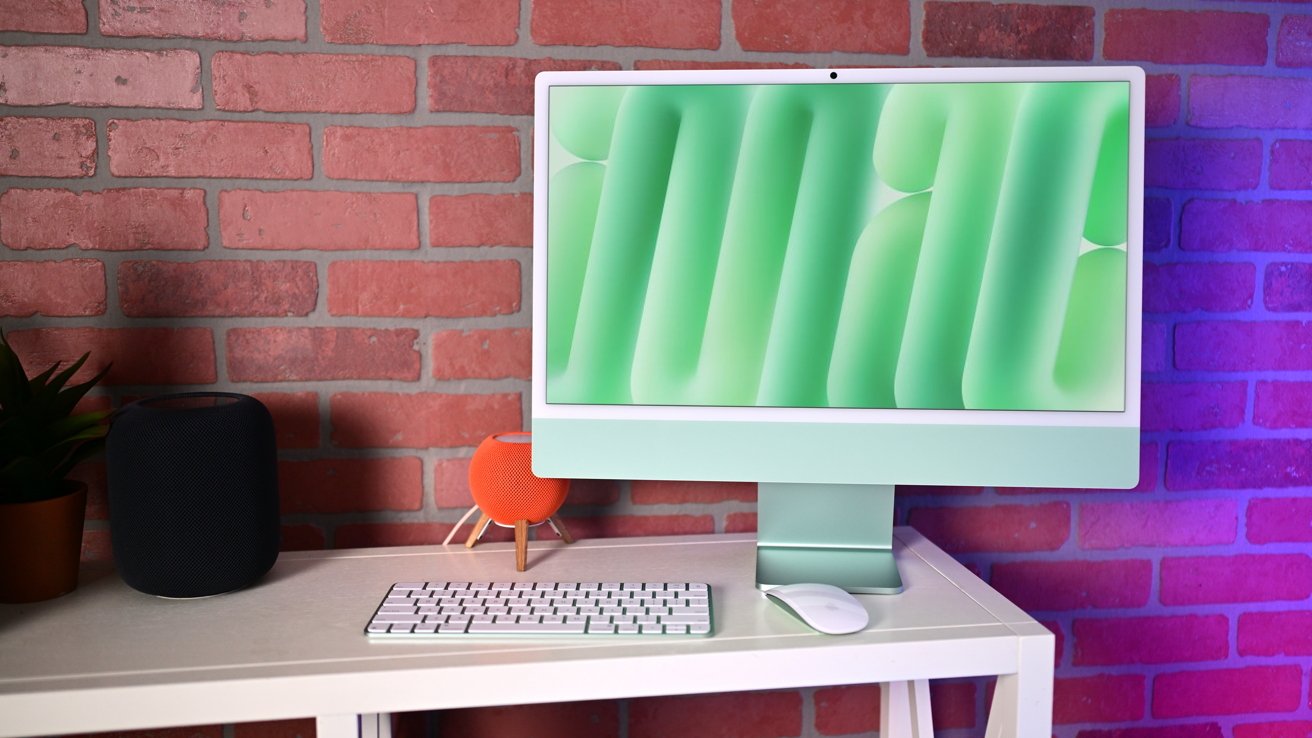 A sleek computer setup with a green-themed monitor, keyboard, mouse, black speaker, and small orange speaker on a white desk against a brick wall.