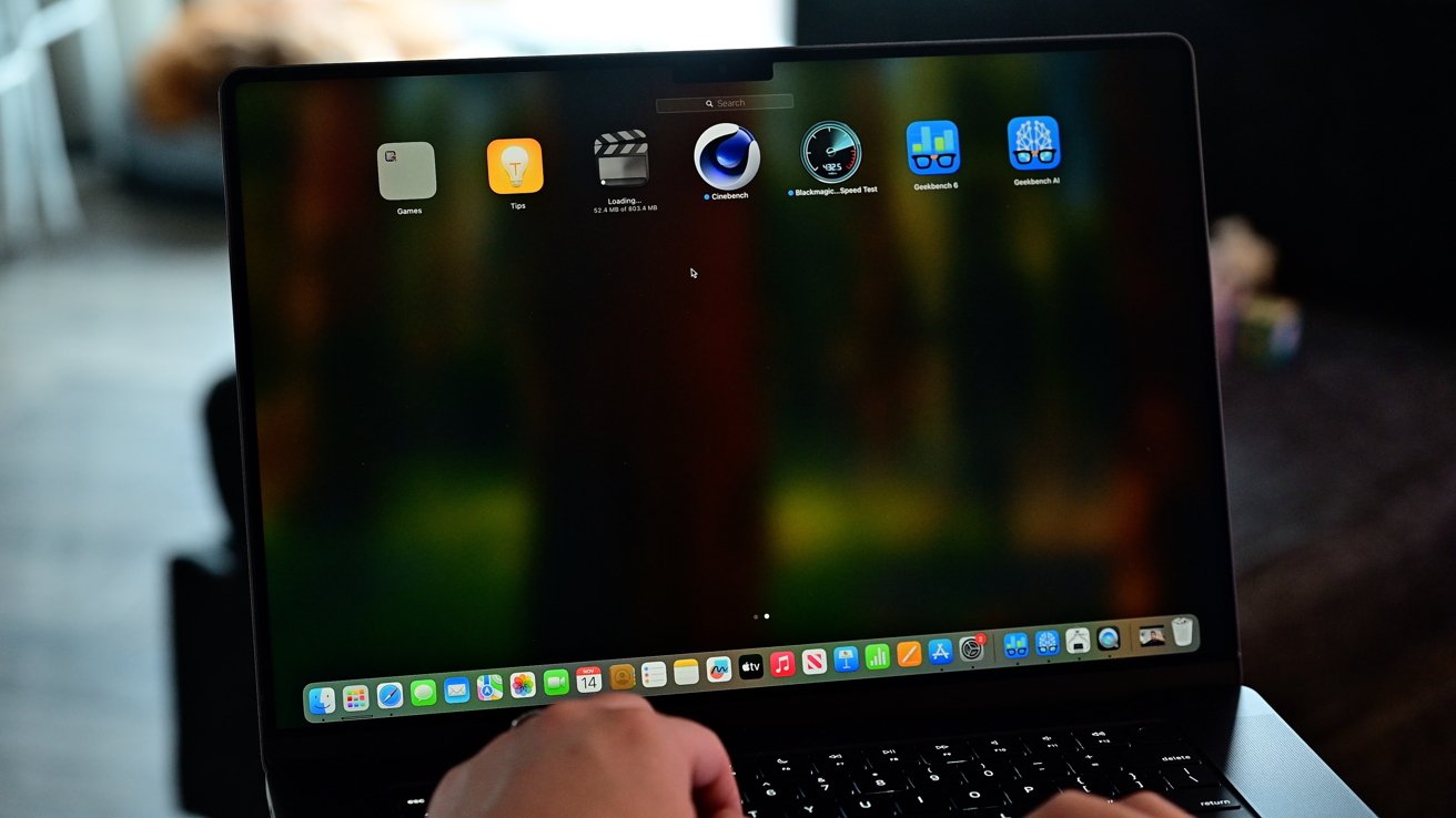 Laptop screen with various app icons, dark reflections visible, and a person typing on the keyboard.