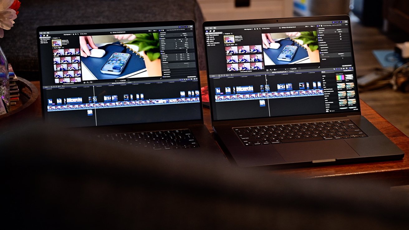 Two laptops displaying video editing software on a wooden table, with a dark blurred object in the foreground.