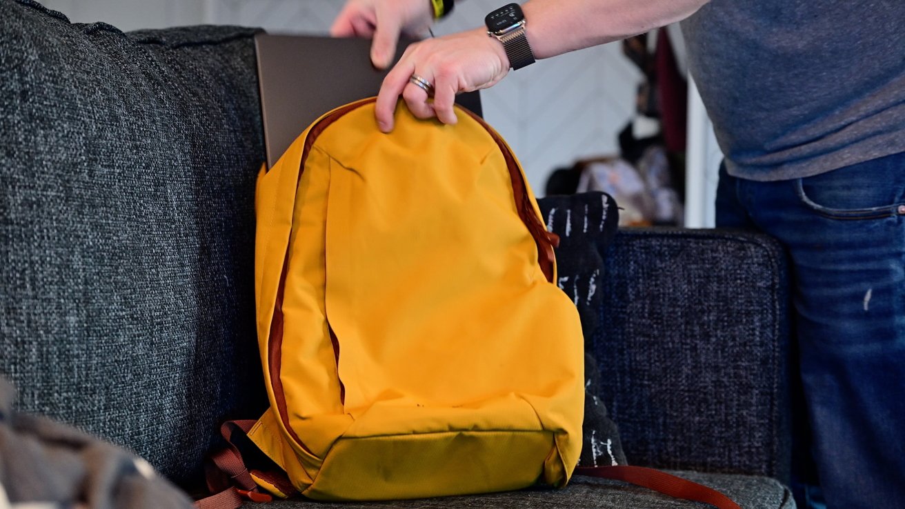 Person placing a laptop into a yellow backpack on a gray couch, wearing a smartwatch and jeans.