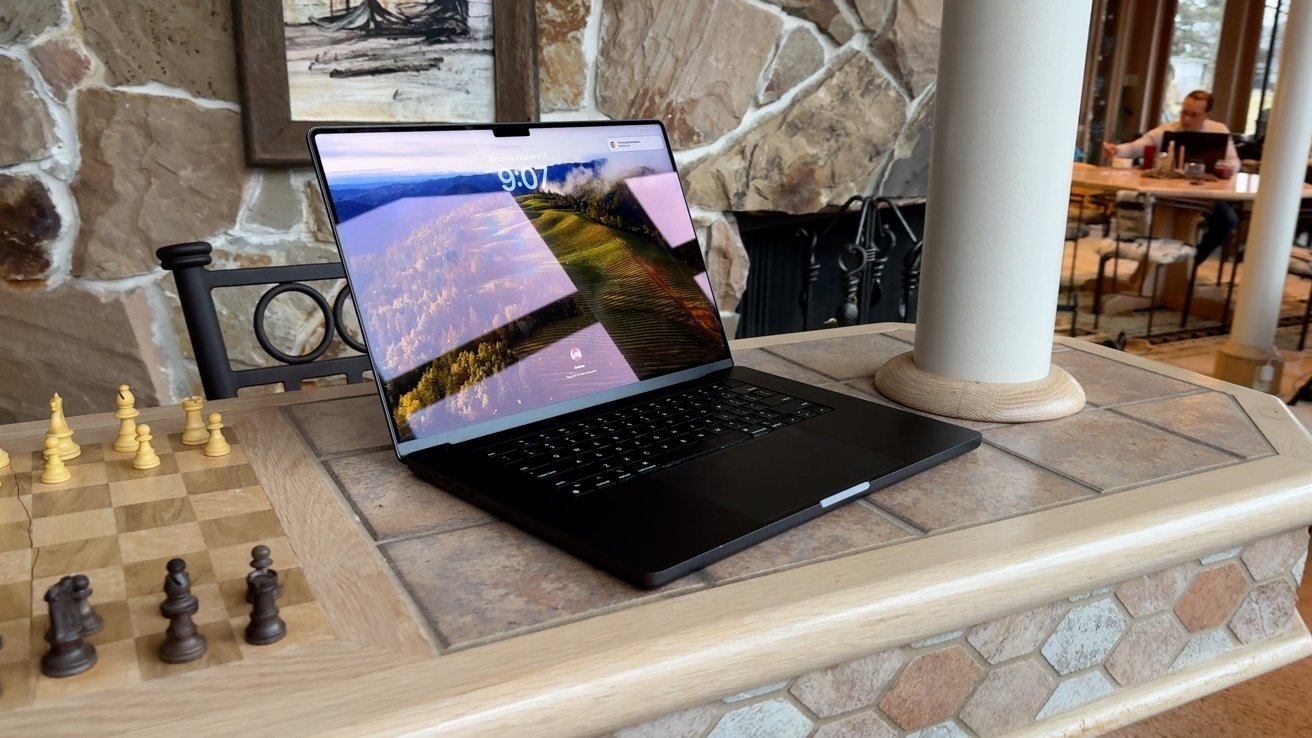 A 16-inch MacBook Pro displaying a scenic landscape photo sits on a stone counter near a chessboard with a stone fireplace in the background.