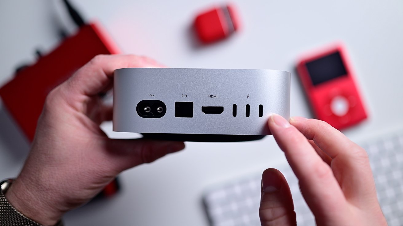 Hands holding a small silver computer device with multiple ports, including HDMI and USB-C, against a blurred background with red gadgets.