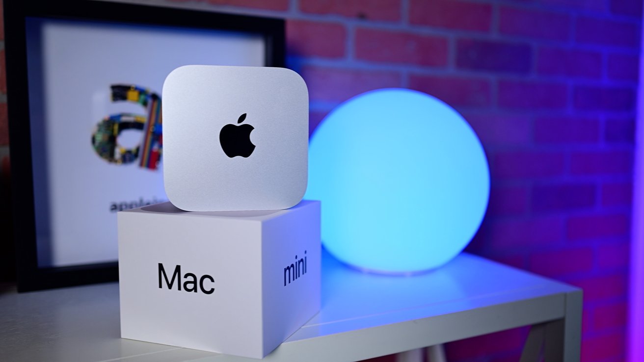 A Mac Mini on a white box labeled 'Mac mini' with a glowing blue sphere and a framed picture in the background.