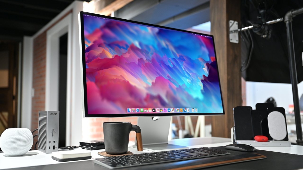 A sleek computer setup with a large Apple Studio Display, black keyboard, coffee mug, and accessories on a modern white desk.