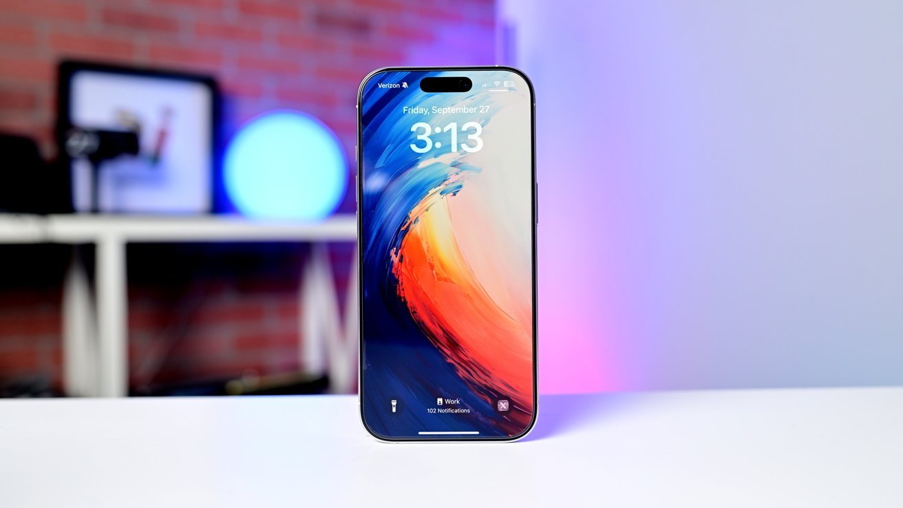 iPhone 16 with vibrant abstract wallpaper displayed on a white surface, with a blurred background featuring a glowing circular light and brick wall.