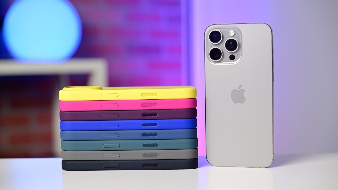 Apple iPhone 16 Pro stands beside a stack of colorful Apple Silicone phone cases, including yellow, pink, red, and shades of blue and green. Background has a blue and purple glow.