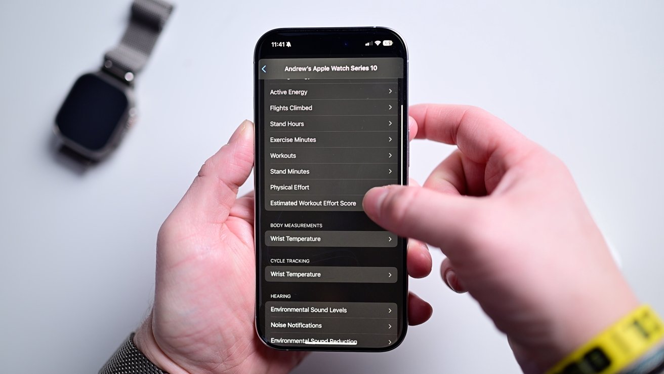 Person holding a smartphone displaying Apple Watch statistics, with a smartwatch on a table in the background.