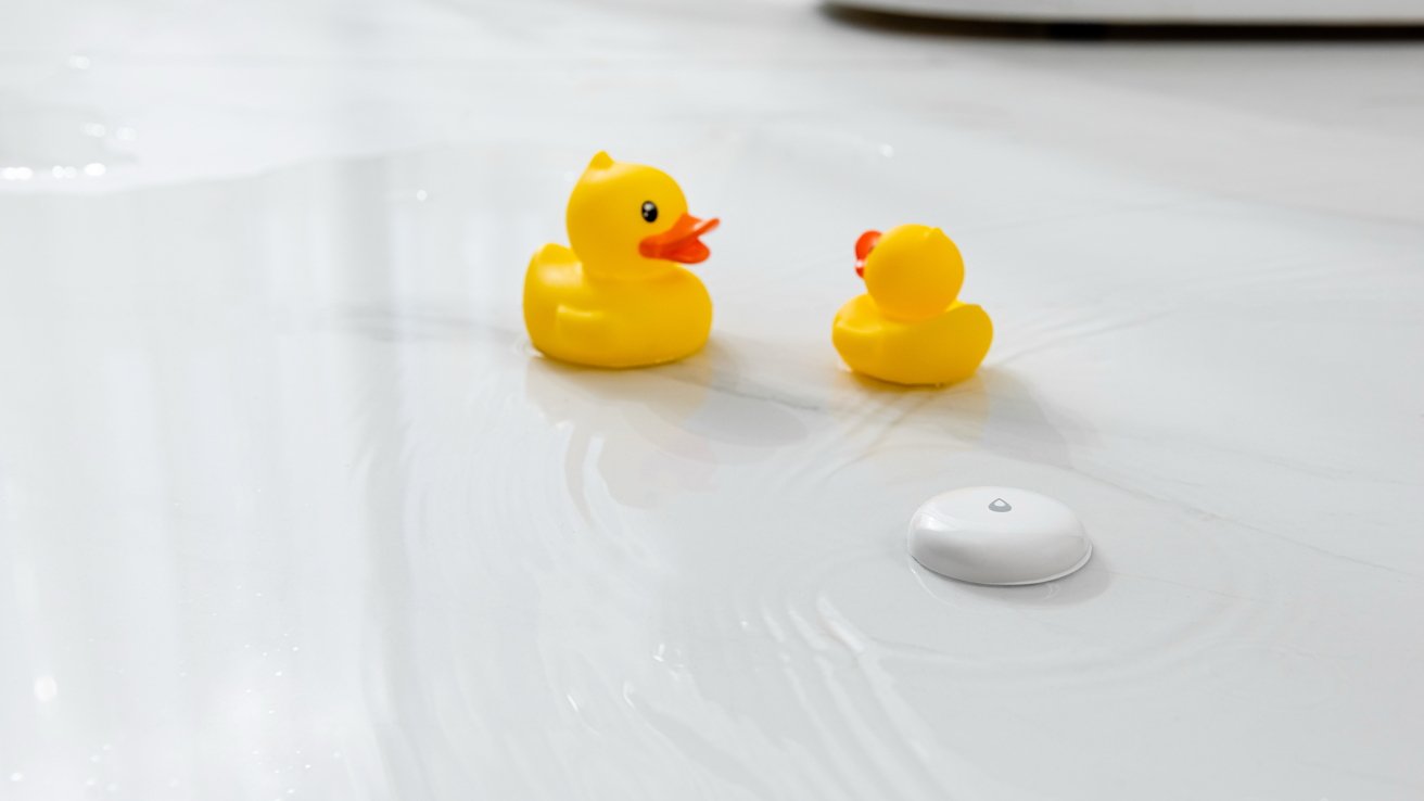 Two yellow rubber ducks on a wet white surface, next to a small white circular device with a water droplet symbol.