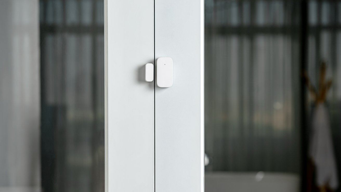 White door sensor on glass door in a room with blurred curtains and a wooden coat rack in the background.