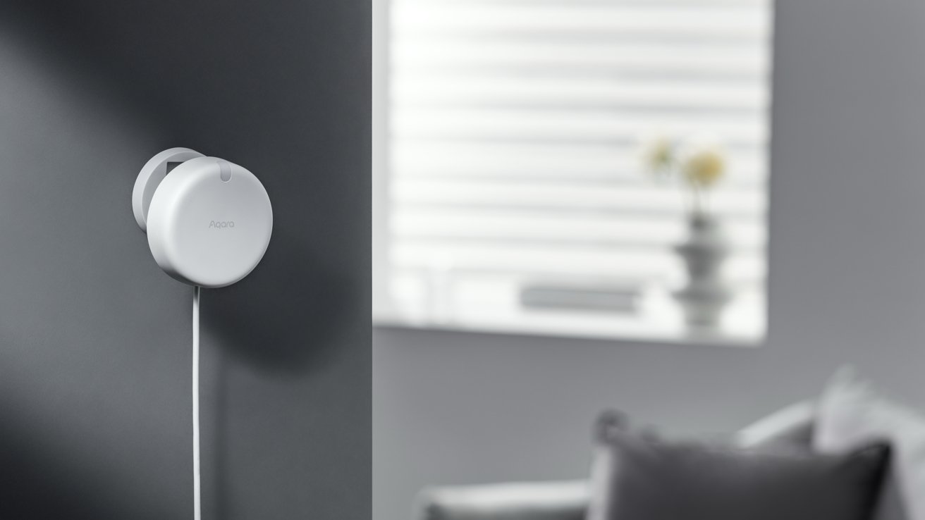 A white circular smart home device mounted on a gray wall with a cord, blurred background with window blinds and sofa.