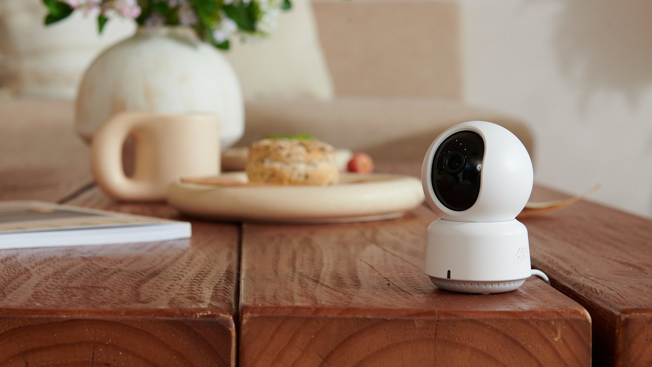 A small white security camera on a wooden table, with a coffee mug and pastry in the background.