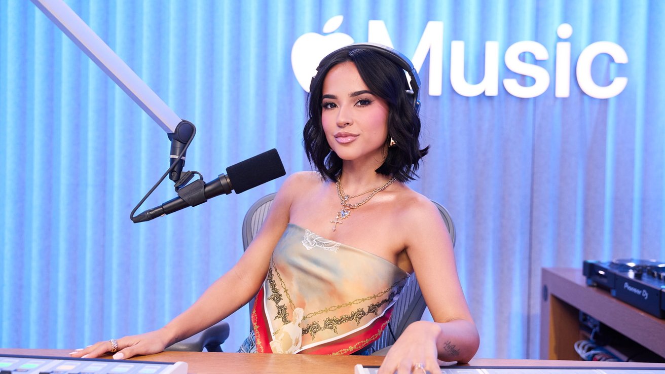 Person sitting at a radio station with headphones, a microphone, and an Apple Music logo in the background, wearing a colorful strapless top.