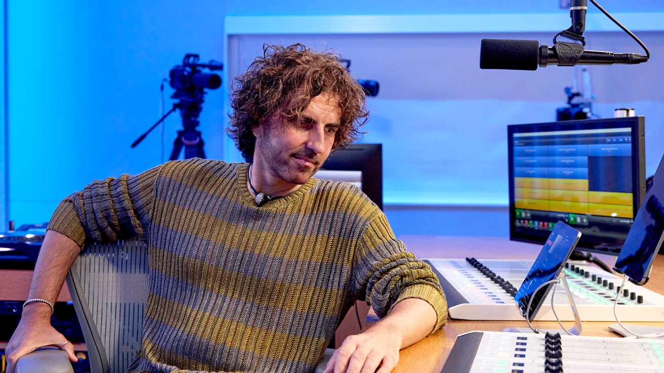 Man with curly hair in a recording studio, wearing a striped sweater, seated by microphones and sound equipment, with multiple screens and cameras in the background.