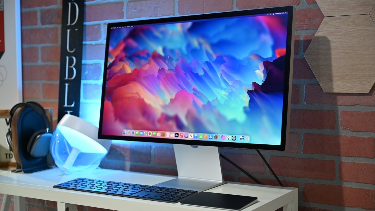 Desktop computer setup with a monitor displaying a vibrant abstract wallpaper, brick wall background, headphones, lamp, keyboard, and trackpad on a white desk.